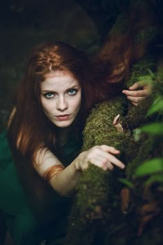 a woman with red hair and blue eyes is leaning on a mossy branch