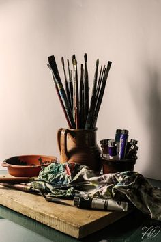 an assortment of art supplies sitting on a wooden table next to a bowl with paintbrushes in it