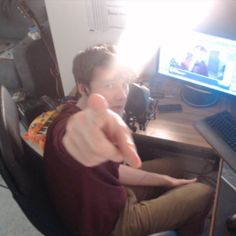 a man sitting in front of a computer desk pointing at the camera with his thumb up