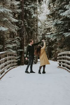 a man and woman standing on a bridge in the snow with trees behind them holding hands