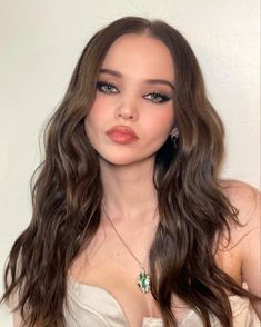 a woman with long brown hair wearing a white top and green earrings, posing for the camera