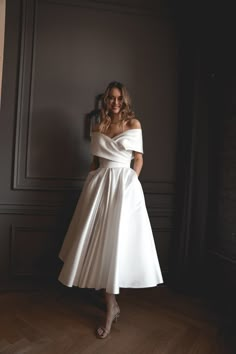 a woman wearing a white dress standing in front of a dark wall with her hands on her hips