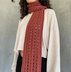 a woman wearing a red crochet scarf and white top standing in front of a gray wall