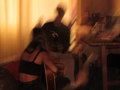 two people sitting on a couch playing guitar in a room with yellow curtains and light coming through the window