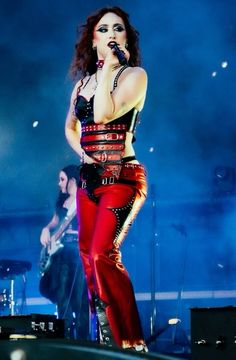 a woman in red pants and black top on stage with microphone, guitar and band behind her