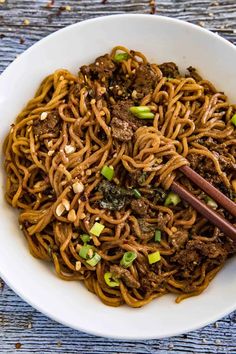 a white bowl filled with beef and noodles on top of a wooden table next to chopsticks