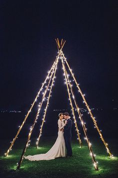 a bride and groom standing in front of a teepee at night