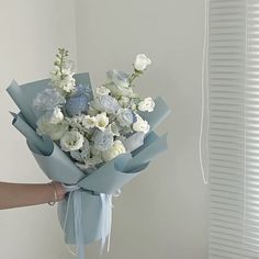 a person holding a bouquet of white and blue flowers in their hand, with blinds behind them