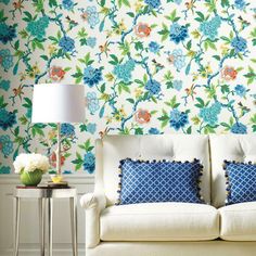 a white couch sitting next to a lamp on top of a table in front of a floral wallpaper