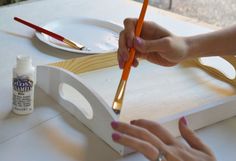 a person holding a pencil in their right hand and painting on a wooden tray with white paint