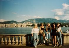 four young women are posing on a bridge near the water and mountains in the background