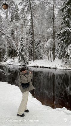 a man standing in the snow taking a photo with his cell phone near a river