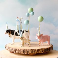 three farm animals with party hats and balloons on top of a tree stump in front of a blue sky