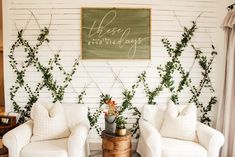 two white chairs sitting next to each other in front of a wall with greenery on it