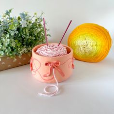 yarn and knitting needles in a pink container next to a plant