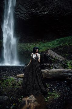 a woman standing in front of a waterfall wearing a long black dress with white lace