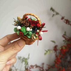 someone is holding a miniature basket with flowers and berries in it on a table next to a plant