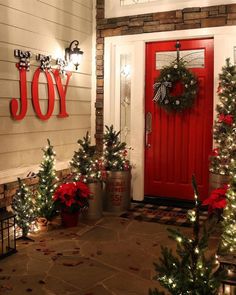 a red door with christmas decorations and lights on the front porch, surrounded by potted trees