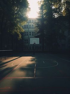 an empty basketball court in the middle of a park with sun shining through the trees