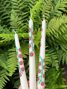 two white candles with red berries on them in front of green leaves and fern plants