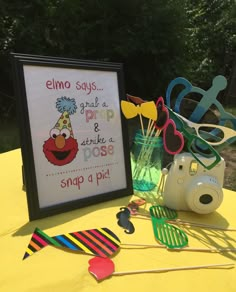 a yellow table topped with glasses and pictures next to a sign that says, elmo says grab a pop & strike a pose snap a pic