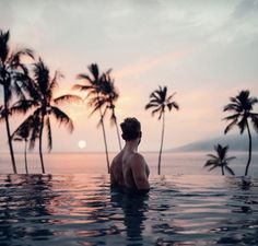 a man is sitting in the water with palm trees around him and looking at the sunset