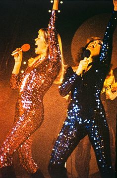 two women in sequin outfits on stage with one holding up her hand to the sky