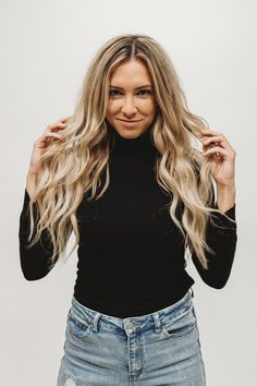 a woman with long blonde hair posing for the camera, wearing ripped jeans and a black turtle neck top