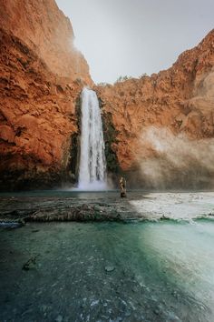 a man standing in front of a waterfall on top of a body of water next to a rocky cliff