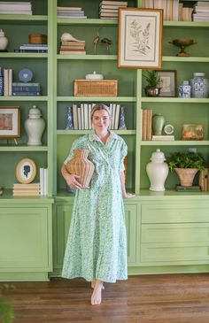 a woman standing in front of green bookshelves holding a basket and wearing a dress