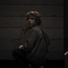 a young man sitting on the ground in front of a wooden wall and wearing a hoodie