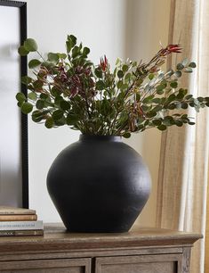 a black vase filled with green plants on top of a wooden table next to a mirror