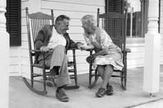 an old man and woman sitting on rocking chairs talking to each other in front of a house