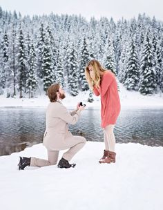 a man kneeling down next to a woman in the snow near a lake and pine trees