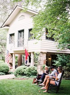 four people sitting on a bench in front of a house with an american flag hanging from the porch