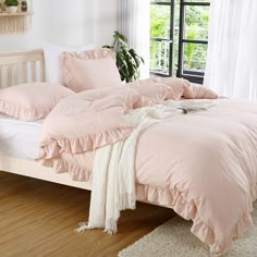 a bed with pink sheets and pillows in a bedroom next to a large window on a wooden floor