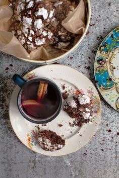 a cup of coffee next to a plate with some cookies on it and a bowl full of powdered sugar