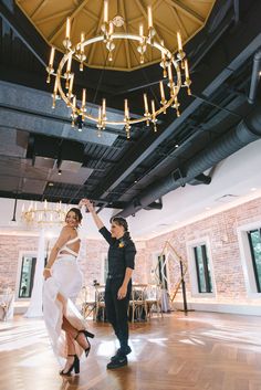 a man standing next to a woman on top of a hard wood floor under a chandelier