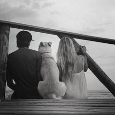 a man and woman sitting on a dock with a dog looking out at the ocean