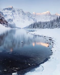 a lake surrounded by snow covered mountains with trees in the foreground and ice on the ground