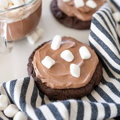 cookies with marshmallows and chocolate frosting are on a striped towel next to a cup of hot cocoa