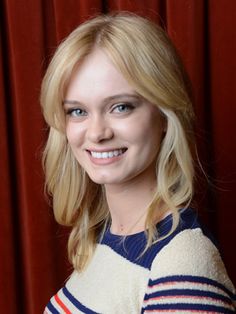 a woman with blonde hair and blue eyes smiles at the camera while standing in front of a red curtain