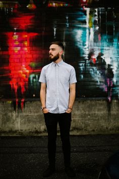 a man standing in front of a wall with graffiti on it and looking off to the side