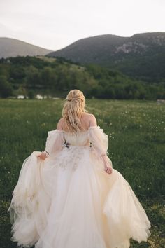 a woman in a white dress is walking through the grass with mountains in the background