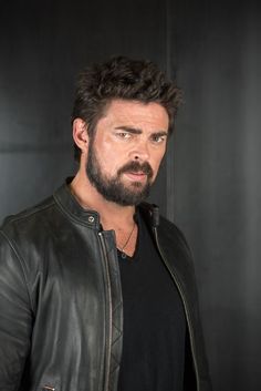 a man with a beard wearing a black shirt and leather jacket standing in front of a wall