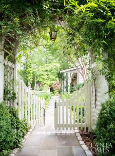 a white picket fence with trees and bushes around it, leading to a small house