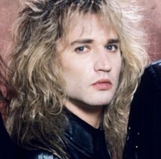 a man with long blonde hair wearing a black leather jacket and looking at the camera