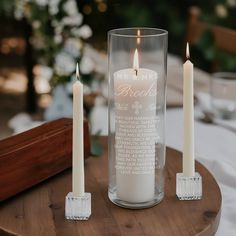 three white candles sitting on top of a wooden table next to a glass vase with writing