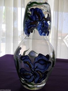 a glass vase with blue flowers in it sitting on a purple table cloth next to a window