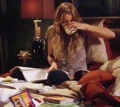 a woman drinking from a glass while sitting in bed with books and magazines on the floor